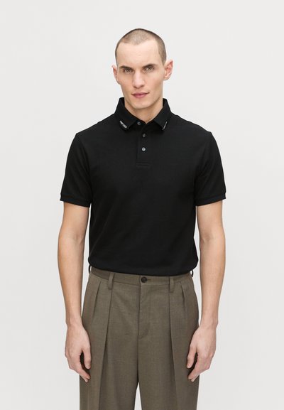 Black polo shirt with a collar, two-button placket, and logo detail on the collar. Slim fit, paired with taupe pleated trousers.