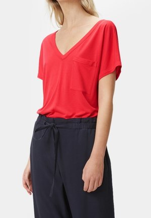 Frau trägt rotes V-Ausschnitt Kurzarmshirt mit Brusttasche und schwarze Hose mit Kordelzug, steht vor einfarbigem Hintergrund.