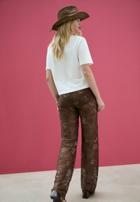 White short-sleeved top paired with brown sheer floral-embroidered pants, featuring a wide-leg cut and patterned texture. Brown straw hat accent.