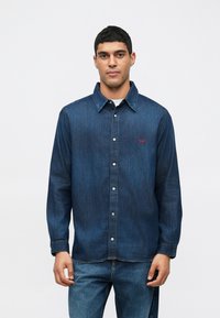 Άνδρας φορώντας ένα μακρυμάνικο σκούρο μπλε τζιν πουκάμισο με κουμπιά και μπλε τζιν, στέκεται μπροστά σε λευκό φόντο.