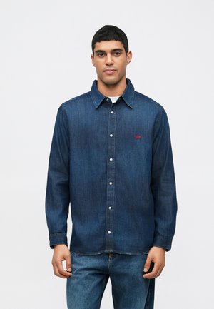 Mies, joka on pukeutunut pitkähiharaiseen tummansiniseen farkkupaitaan, jossa on napit, ja sinisiin farkkuihin, seisoo yksinkertaista valkoista taustaa vasten.