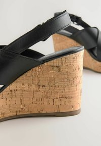 Sandali con zeppa in pelle nera e tacco in sughero. Il tacco presenta una superficie in sughero texturizzato, e le cinghie si incrociano sulla parte superiore del piede.