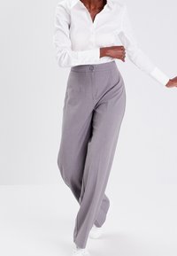 Pantalon gris taille haute avec plis à l'avant, fermeture par boutons, coupe large et texture lisse, associé à une chemise blanche à boutons.