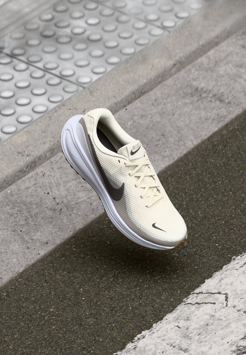 Nike trkaće cipele, bež mrežasti gornji deo, teksturirana površina, crni Swoosh, bijela potplat, aerodinamičan dizajn, sistem vezivanja, lagana konstrukcija.