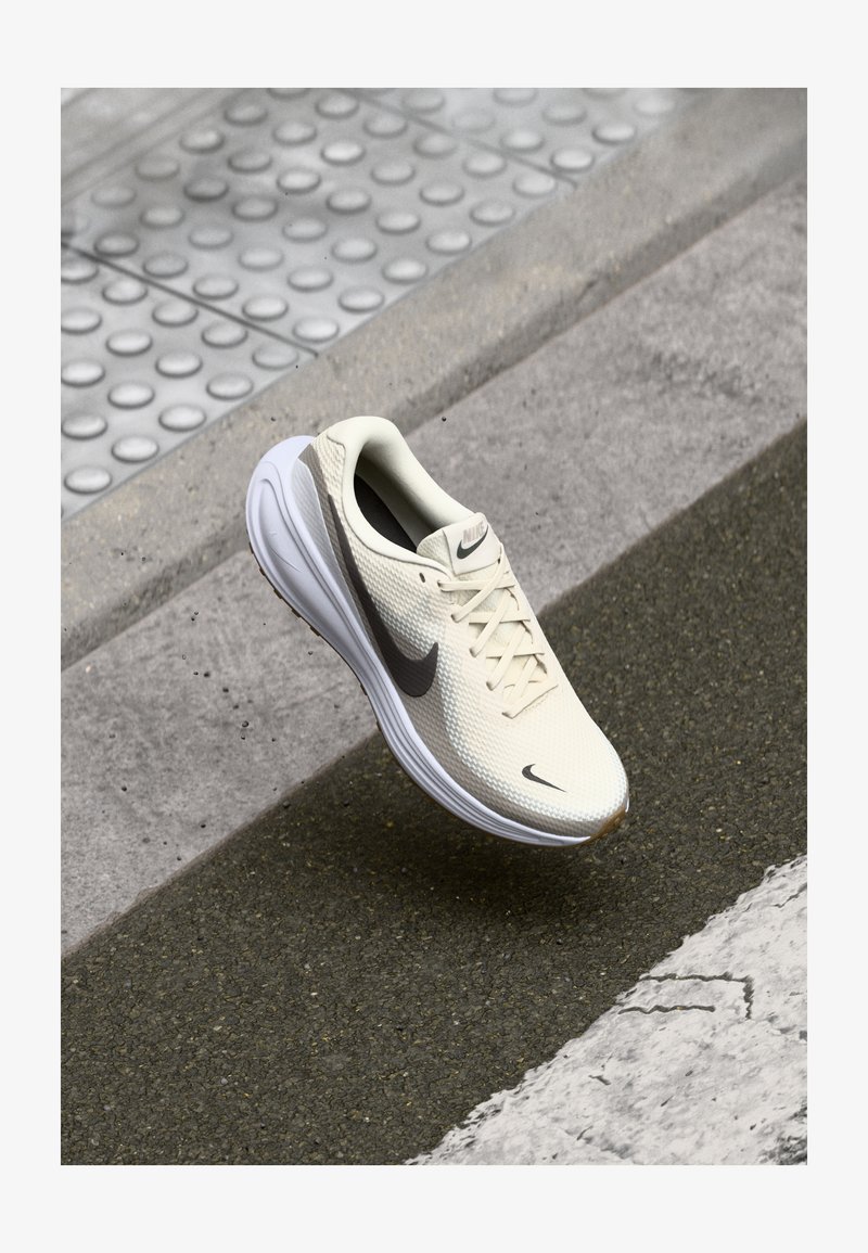 Nike trkaće cipele, bež mrežasti gornji deo, teksturirana površina, crni Swoosh, bijela potplat, aerodinamičan dizajn, sistem vezivanja, lagana konstrukcija.
