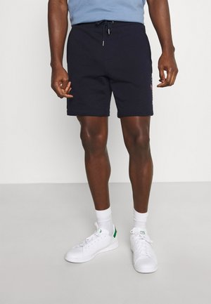 Mand iført sorte shorts med snor, hvide sokker og hvide sneakers, står på en lys grå gulv mod en hvid væg.