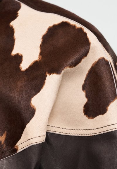 Detailný záber na vzor hnedo-bielej hovädzej kože na prešívanom koženom povrchu, zobrazujúci textúru a prirodzené detaily srsti.