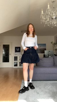 Mlada ženska stoji v notranjosti oblečena v belo majico z dolgimi rokavi, črno nabirno jeans krilo, bele nogavice in črne platforme.