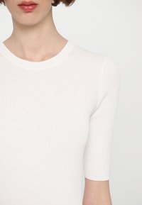 Haut blanc à manches courtes côtelé avec un col rond. Le tissu semble doux avec une texture subtile. Pas de motifs ou d'accents notables.
