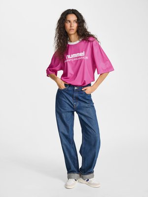 Kvinde med krøllet hår iført en knaldrød Hummel Performance trøje, løse blå jeans og hvide sneakers, stående med hænderne i lommerne.