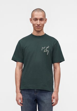 Homme à la tête rasée portant un t-shirt vert foncé avec une inscription blanche "just get lucky" sur la poitrine et un jean bleu, debout devant un fond uni.