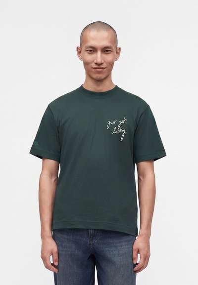 Homme à la tête rasée portant un t-shirt vert foncé avec une inscription blanche "just get lucky" sur la poitrine et un jean bleu, debout devant un fond uni.