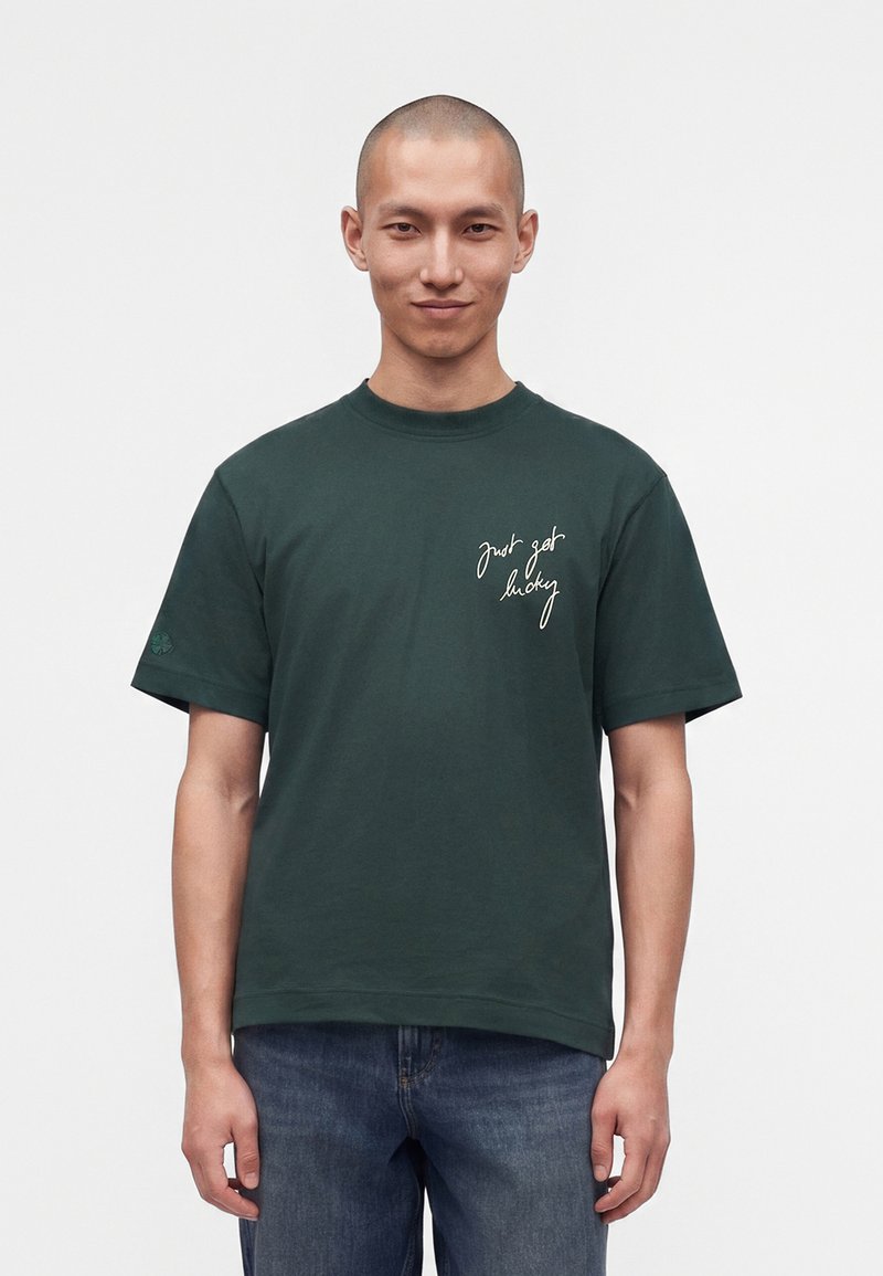 Homme à la tête rasée portant un t-shirt vert foncé avec une inscription blanche "just get lucky" sur la poitrine et un jean bleu, debout devant un fond uni.