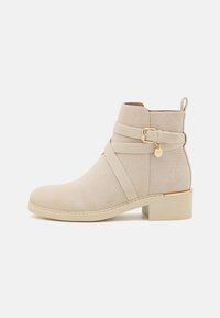 Bottines beiges en daim, avec un design à croisillons, un accent de boucle dorée et un dos texturé. Talon épais et bas.