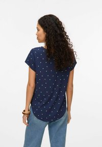 Marineblauwe top met korte mouwen en ronde zoom, gekenmerkt door kleine groene stippen in een verspreid patroon. De stof lijkt lichtgewicht en glad.