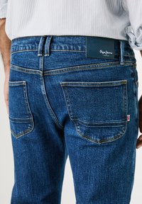Sinivärvi denim püksid, millel on nahast vöötael, millel on "Pepe Jeans" logo. Kontrastne õmblemine, kaks tagumist taskut, millel on kõverad servad.