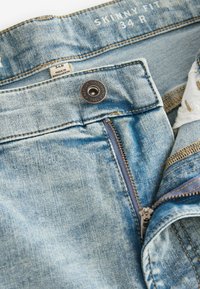 Svijetloplave traperice uskog kroja, s zatvaračem na zip i brončanim gumbom. Uključuje oznaku s informacijama o veličini i podrijetlu.