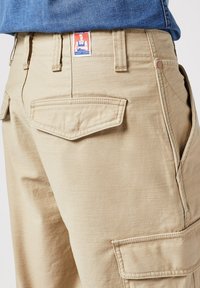 Pantaloni cargo beige con tasche doppio cucite, cuciture rinforzate e un'etichetta tessuta in vita, dotati di zip e passanti per cintura.