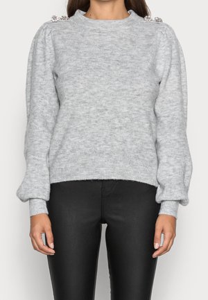Pull-over en maille gris clair avec manches longues bouffantes et boutons décoratifs en forme de bijoux sur l'épaule, porté avec un pantalon noir ajusté.