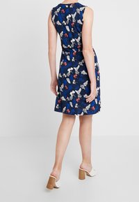 Robe florale avec un fond marine, ornée de fleurs rouges et blanches, sans manches et avec un ourlet évasé. Associée à des mules blanches à talons.