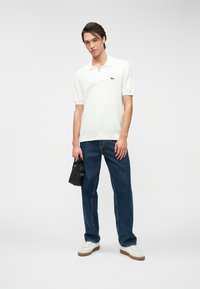 Polo blanc pour homme à manches courtes, en tissu texturé, associé à un jean bleu marine et des baskets blanches. Tenant un petit sac à main noir.