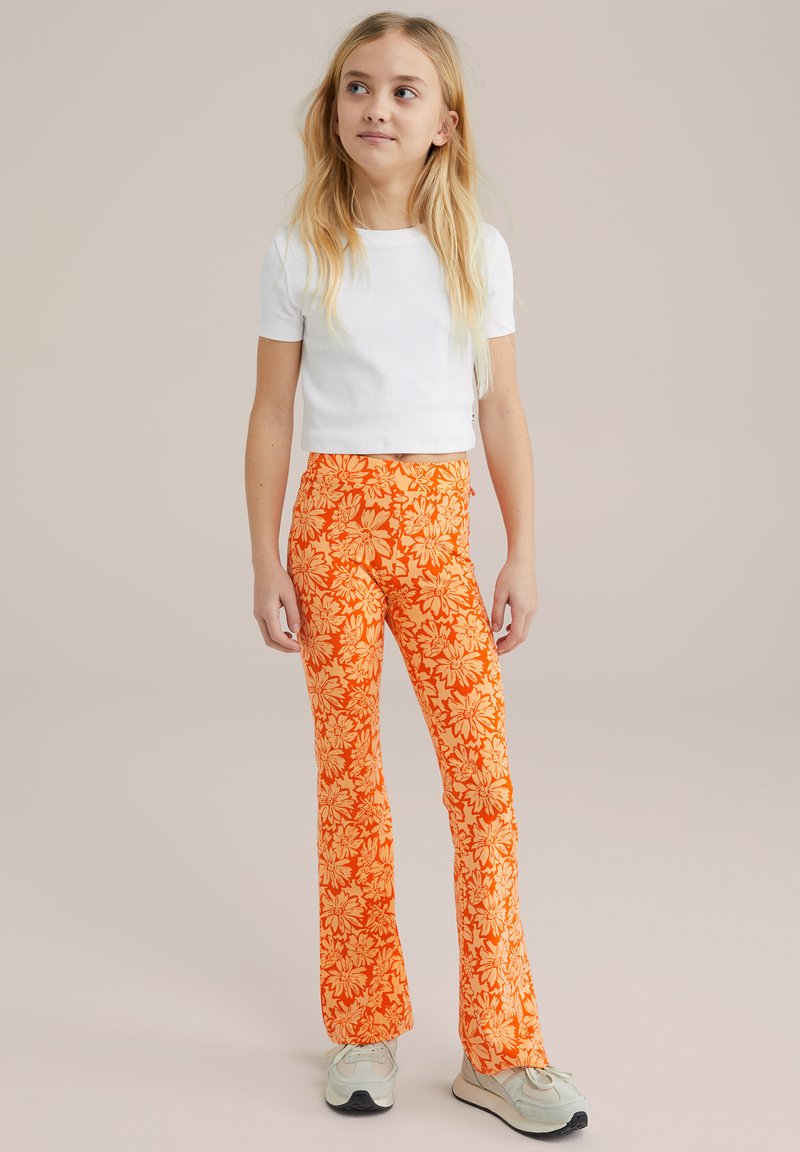 Rdeče hlače z razširjenimi hlačnicami in živahnim cvetličnim vzorcem, v kombinaciji z belo kratko majico s kratkimi rokavi. Obutev so svetlo sivi športniki.