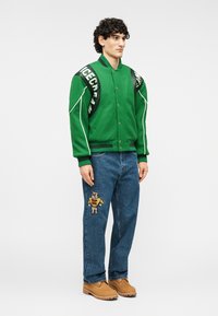 Πράσινο αθλητικό μπουφάν με άσπρες λεπτομέρειες, μπλε τζιν παντελόνι με κεντημένη αρκούδα και καφέ παπούτσια. Συμπληρωματικά αξεσουάρ: λουρί σακιδίου πλάτης με επώνυμο λογότυπο.