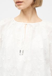 Γυναίκα που φορά λευκό διάφανο μπλουζάκι με κεντήματα λουλουδιών και ντεκολτέ με δέσιμο μπροστά με κορδόνια με ασημένιες άκρες.