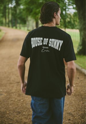 Junger Mann mit dunklem Haar, der auf einem unbefestigten Weg in einem Park geht, trägt ein schwarzes T-Shirt mit der Aufschrift "HOUSE OF SUNNY Hackney London".