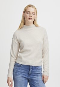 Maglione beige chiaro con colletto alto a coste e maniche lunghe, caratterizzato da una texture morbida e orlo aderente, abbinato a jeans blu.