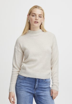 Pull beige clair avec un col haut en côtes et des manches longues, présentant une texture douce et un ourlet ajusté, associé à un jean bleu.