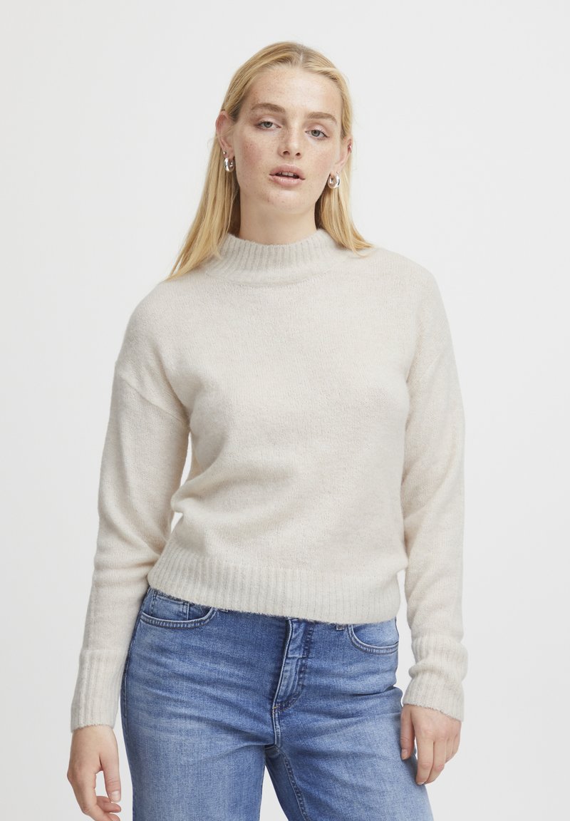 Maglione beige chiaro con colletto alto a coste e maniche lunghe, caratterizzato da una texture morbida e orlo aderente, abbinato a jeans blu.