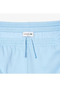 Svetlo modre usnjene trenirke s elastičnim pasom, opremljene z vrvico in oznako blagovne znamke Lacoste. Gladka tekstura blaga.