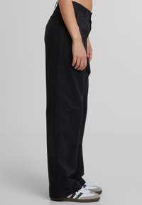 Wide-leg black trousers featuring a classic fit, side pockets, and subtle logo detail above right pocket. Paired with low-top sneakers.