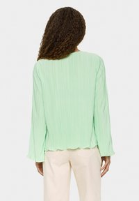 Jasnozielona bluzka z fałdami, z długimi rękawami i zaokrąglonym dołem. Wykonana z teksturowanej tkaniny, prezentująca miękki, swobodny fason.