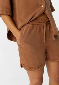 Pantaloni corti in tessuto morbido marrone, con vita elasticizzata e coulisse, dotati di tasche laterali e dettagli di orlo cuciti per una maggiore texture.