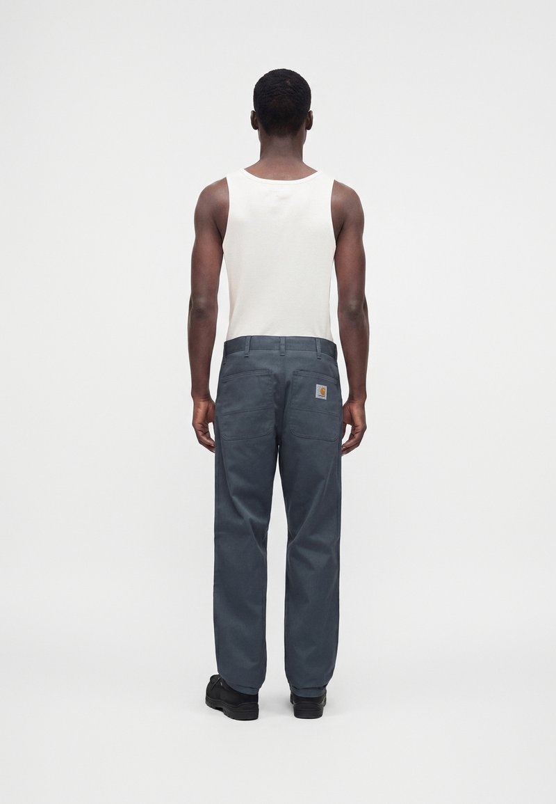 Homme debout face à l'arrière, portant un haut blanc sans manches, un pantalon de travail bleu foncé avec des poches arrière et des chaussures noires sur un fond uni.