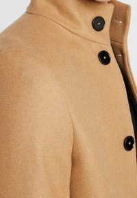 Beige Wollmantel mit hohem Kragen, drei schwarzen Knöpfen und einem strukturierten Stoff. Klare Linien mit taillierter Passform und subtilen Stickdetails.