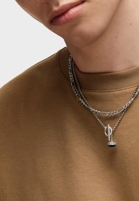 T-shirt in jersey marrone con scollatura rotonda, sovrapposto a due catene d'argento. Una catena presenta un ciondolo con una base circolare e un dettaglio pendente.