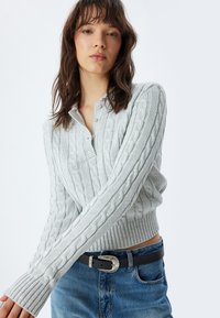 Maglione a collo alto grigio chiaro a maglia cable-knit con orlo e polsini a costine, caratterizzato da un colletto con bottoni e motivi testurizzati. Indossato con jeans denim blu.