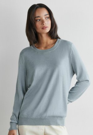 Młoda kobieta o prostych, ciemnych włosach, ubrana w jasno-niebieski sweter z długim rękawem i białe spodnie, stojąca na tle gładkiej białej ściany.