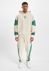 Beige hettegenser og bukser med tealfarget detaljer. Har en logo på brystet og hoften. Laget av myk, teksturert stoff. Hvite sneakers fullfører antrekket.