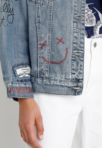 Jeansová bunda s červeným vyšitým usmívajícím se obličejem, text "LA VIDA" na manžetě, nášivka s rty "bonjour", nošena s bílými kalhotami a námořnickou květinovou košilí.