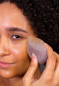 Makeupsvamp med rund form, mjuk textur och gradientfärger från beige till lila, hålls mot en hudtonad bakgrund.