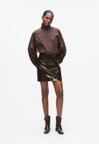 Blusa a collo alto marrone con maniche voluminose, abbinata a una mini gonna lucida marrone scuro a vita alta e stivali alla caviglia coordinati.