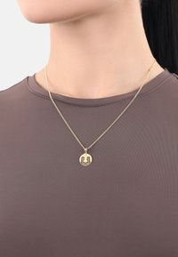 Guldkedja med en cirkulär hänge som har en detaljerad design. Bärs över en enfärgad, åtsittande brun topp. Kedjan är delikat och har en texturerad yta.
