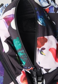 Intérieur de sac à dos en tissu multicolore avec une fermeture éclair noire, des motifs floraux et une étiquette noire texturée, mettant en valeur diverses teintes vives.