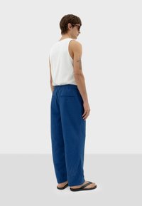 Blauwe textuurbroek met een elastische tailleband, gecombineerd met een witte mouwloze top. De persoon draagt zwarte slippers en heeft een tattoo.