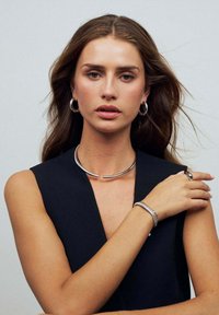 Silverhalsband med en böjd design, hoop-örhängen och ett texturerat armband som bärs över en svart topp. Slät metallfinish, minimalistisk stil.