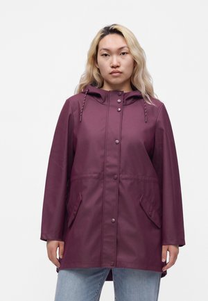 Jeune personne aux longs cheveux blonds portant un imperméable violet à capuche et un jean bleu clair, debout devant un fond uni.